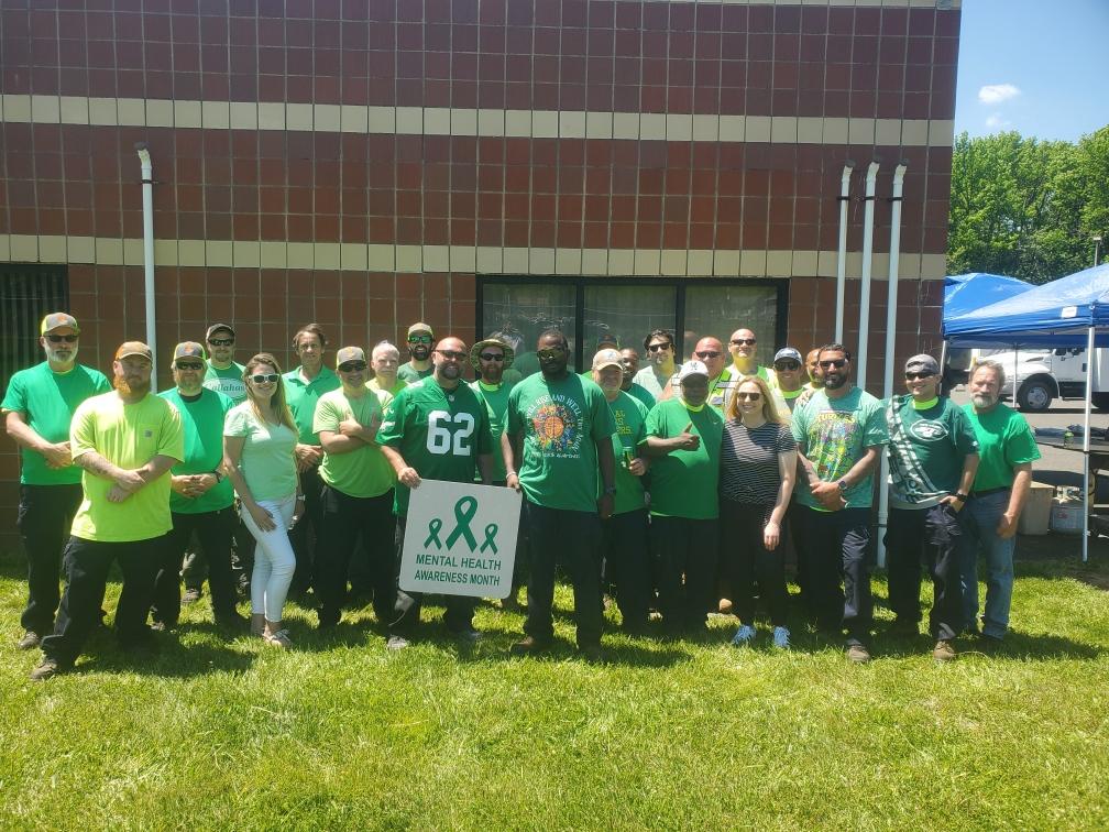Group of volunteers in green shirts posing outdoors in front of a building with a Mental Health Awareness Month banner.